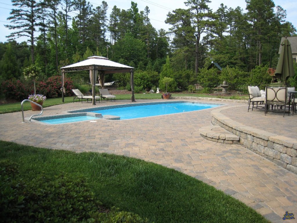 This is a perfect example of what we mean when we say "Outdoor Living Space." Our definition is brilliantly demonstrated by this gorgeous example of an Olympus fiberglass pool - paired perfectly with outdoor accessories. This image shows a nice travertine deck with a raised porch, nice outdoor furniture and natural landscaping to finish it off. This Olympus fiberglass pool looks very natural and serene - a perfect fit for this beautiful home!